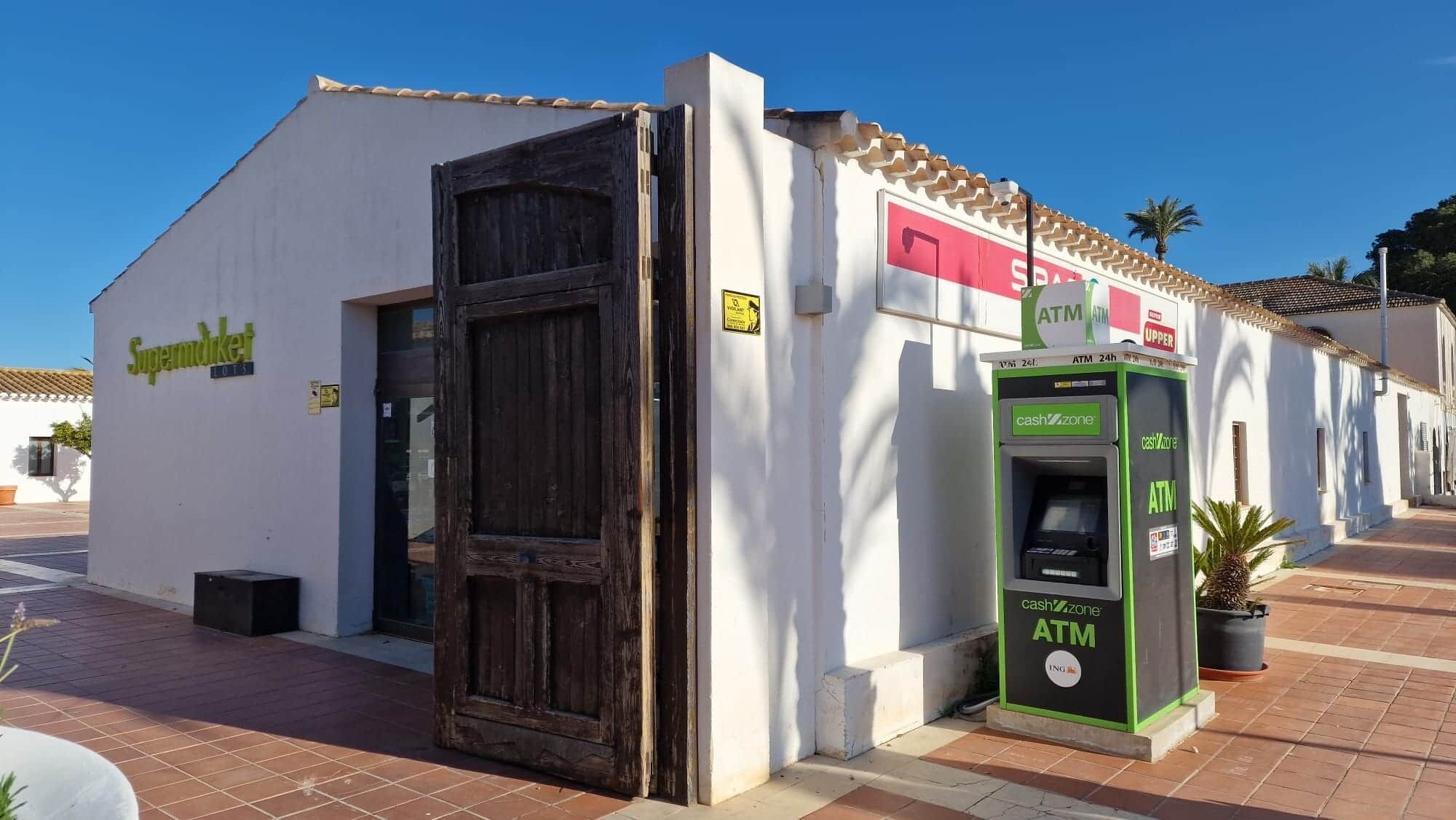 Un bâtiment avec une porte en bois fermée, une enseigne indiquant « Supermarché » et un distributeur automatique de billets étiqueté « zone de retrait d’espèces ».