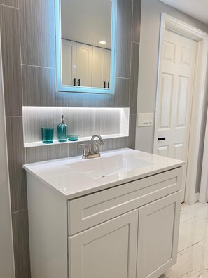 Une salle de bain moderne avec un meuble-lavabo blanc, un grand miroir et une étagère intégrée où sont disposés des flacons en verre.
