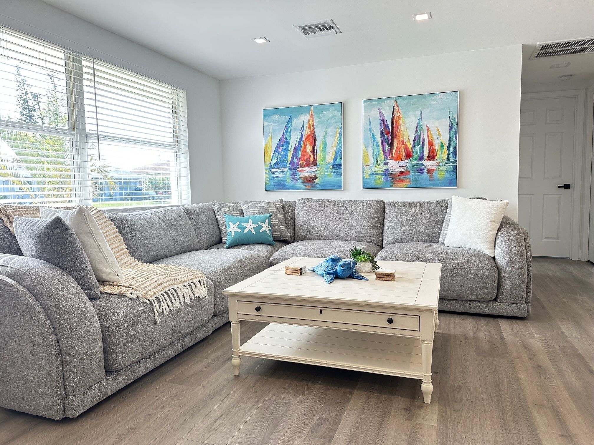 A modern living room with a grey sectional sofa, a white coffee table, and two paintings of sailboats on the wall.