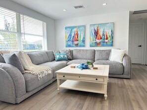 Un salon moderne avec un canapé d’angle gris, une table basse blanche et deux tableaux représentant des voiliers accrochés au mur.