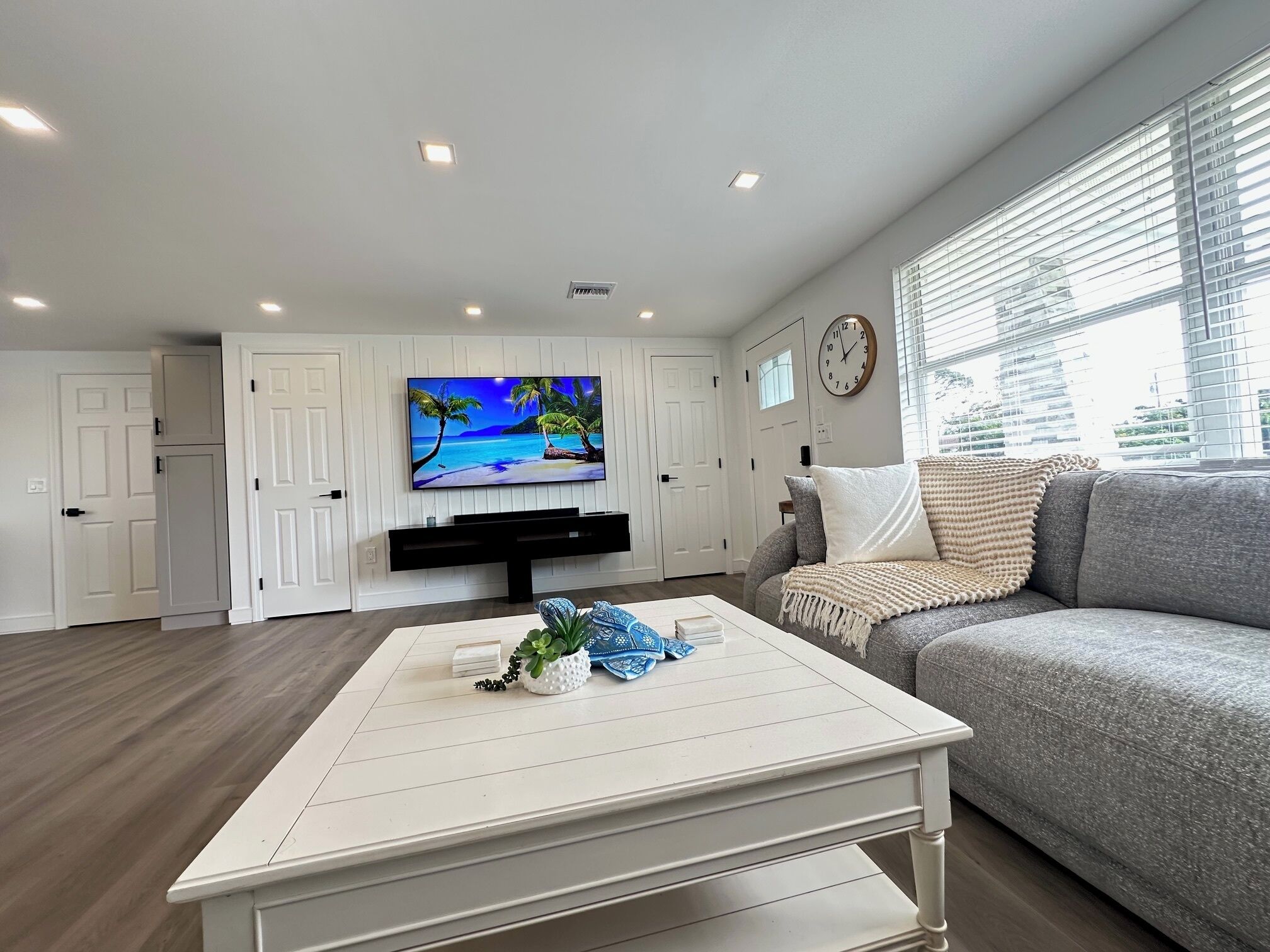 A modern living room with a flat-screen TV mounted on the wall, a grey sofa, a white coffee table with decorative items, and a window with blinds.