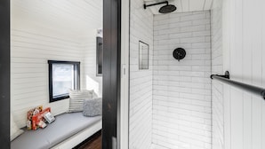 Une salle de bain compacte aux murs recouverts de carreaux blancs, avec une douche équipée d’un pommeau de douche à main, une petite fenêtre et un banc sur lequel sont posés des jeux de Scrabble.