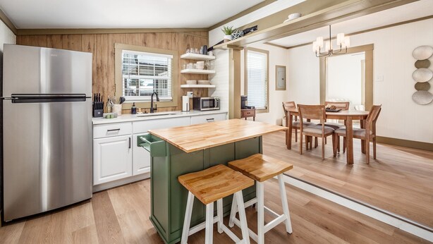A modern kitchen with a stainless steel refrigerator, wooden cabinets, a green bar stool, and a dining area with a wooden table and chairs.