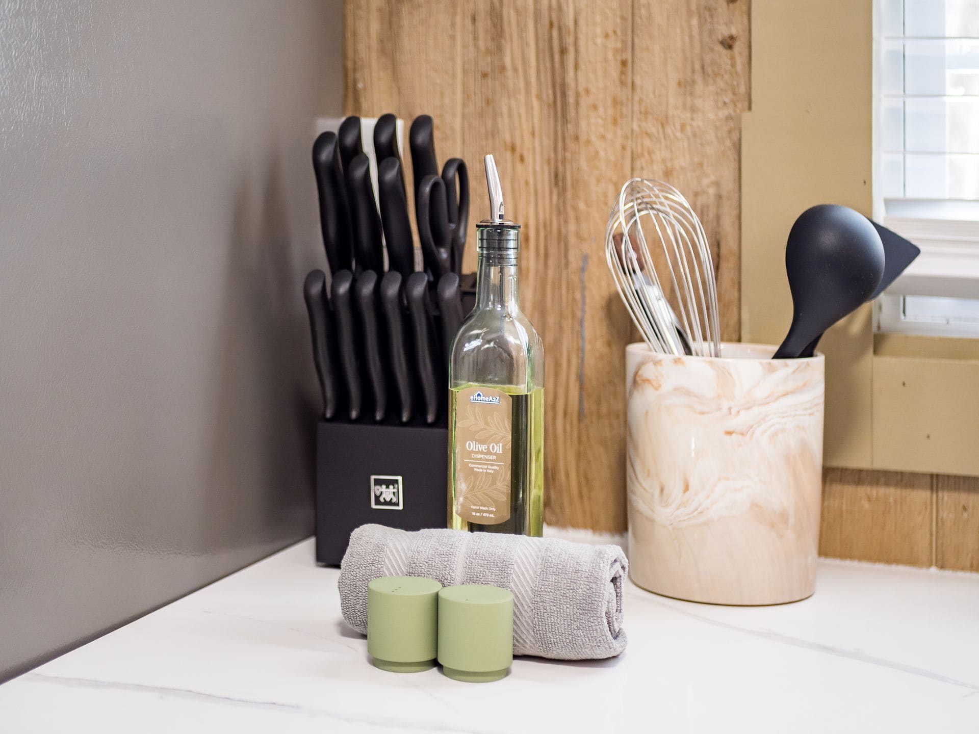 Een keukenblad met een fles olijfolie, een messenblok, een garde, een handdoek en twee kleine bakjes.