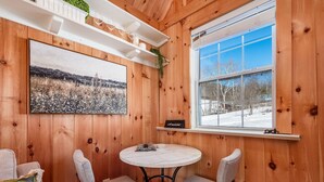 Ein gemütlicher Holzraum mit einem runden Tisch und zwei Stühlen, ein großes Fenster mit Blick auf eine verschneite Landschaft und ein gerahmtes Bild an der Wand.