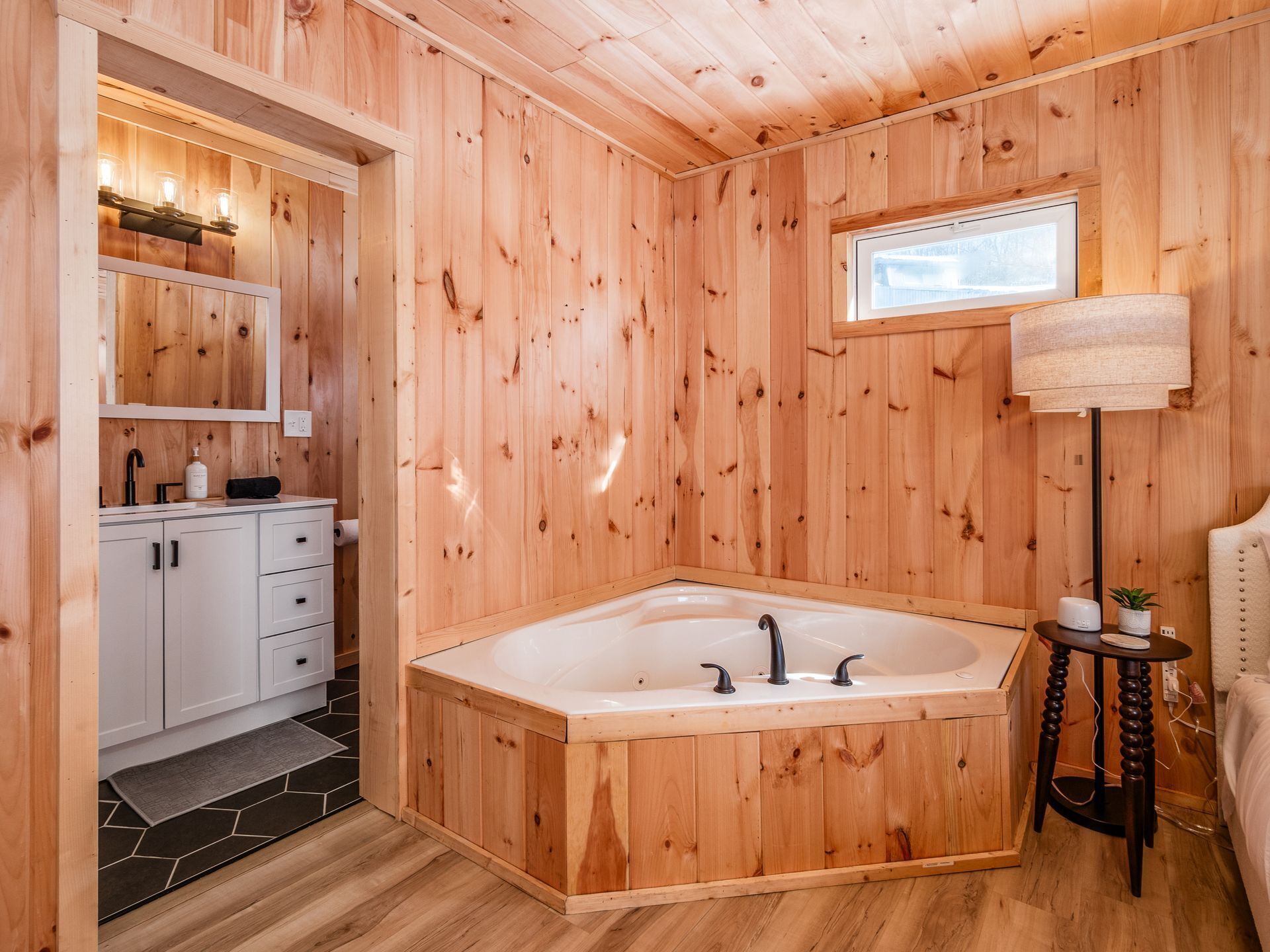A wooden cabin bathroom with a freestanding bathtub, a mirror, a small table with a lamp, and a window.