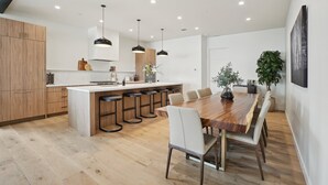 Una cocina moderna con armarios de madera, una isla central con taburetes de bar, iluminación colgante y un comedor con una mesa de madera larga y sillas.
