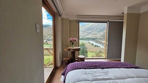 A bedroom with a large window overlooking a valley and river. There is a wooden table with a vase of flowers and a bed with a purple blanket. - Amaru TreeHouse (Cusipata)