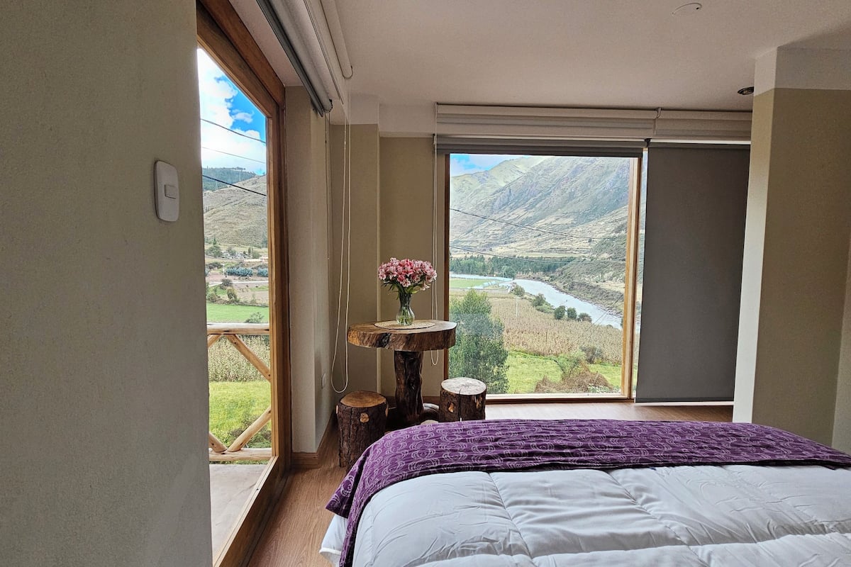 Un dormitorio con un ventanal grande que da a un valle y un río. Hay una mesa de madera con un jarrón de flores y una cama con una manta morada.