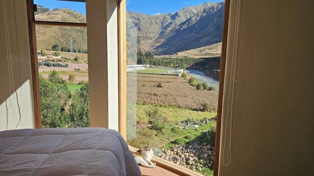 A room with a large window overlooking a mountainous landscape and a river. - Amaru TreeHouse (Cusipata)