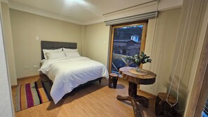 A bedroom with a bed, a small wooden table with a vase, and a view of a building through a window. - Amaru TreeHouse (Cusipata)