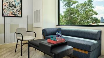 A modern room with a grey sofa, a black bench, and a chair. There is a framed artwork on the wall and a window with a view of trees and buildings.