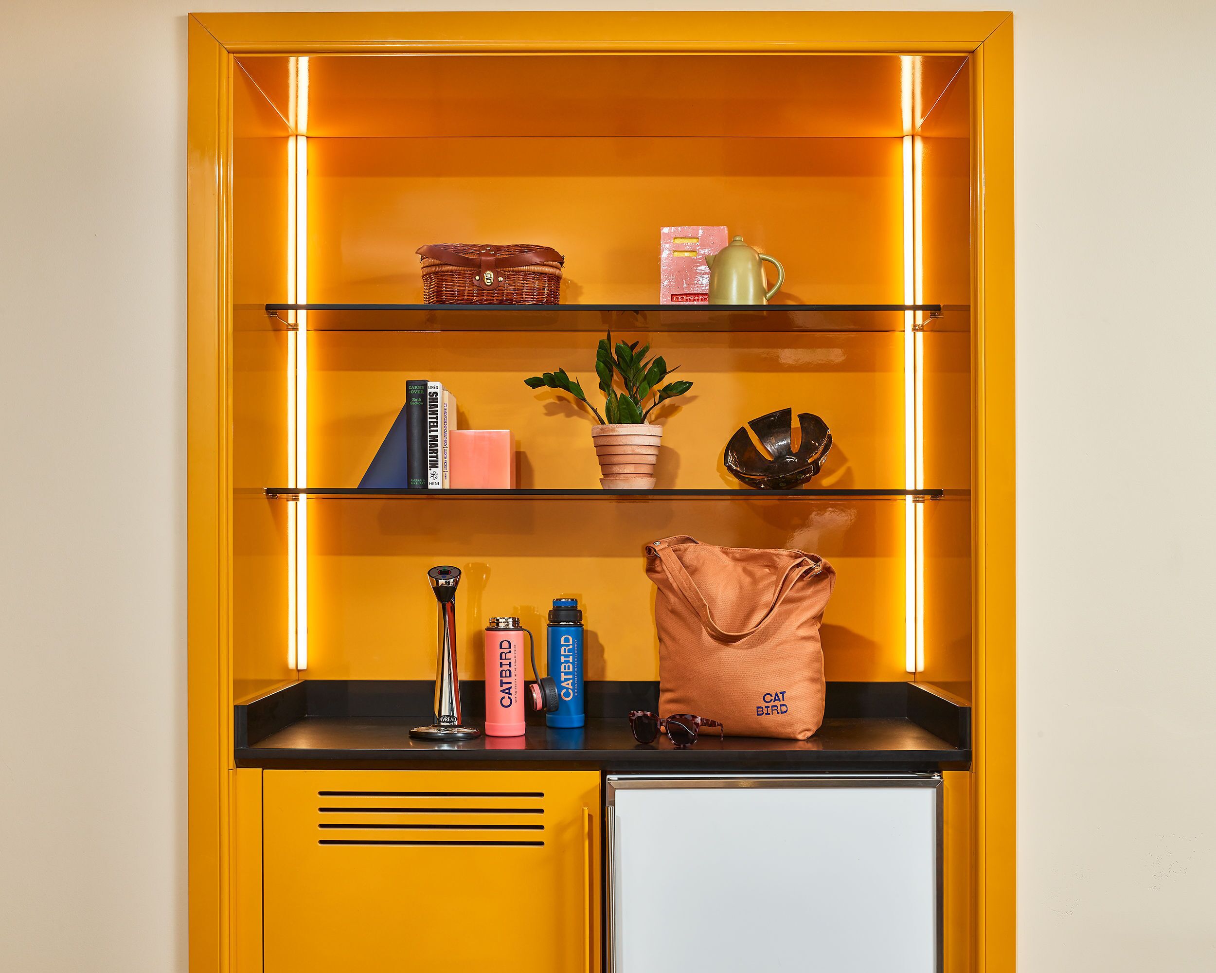 A yellow cabinet with shelves containing books, a plant, a teapot, a handbag, and a small refrigerator.