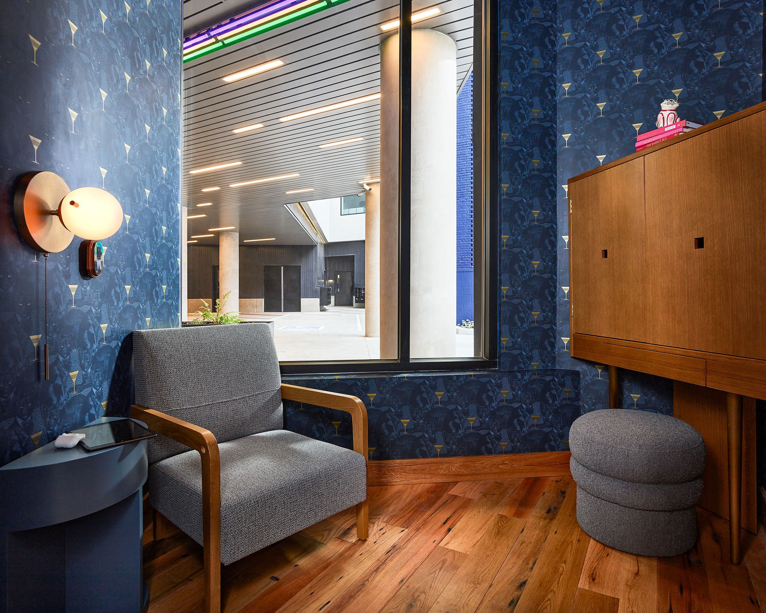 A modern hotel room with a blue patterned wall, a wooden floor, a grey armchair, a small ottoman, and a wooden cabinet.