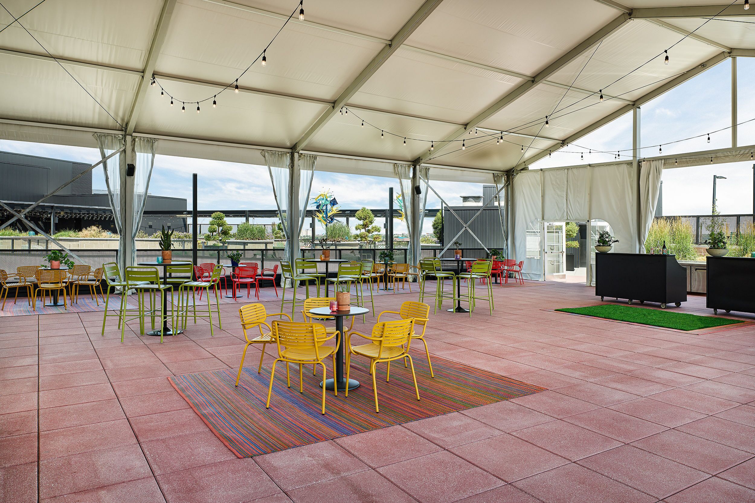 Un espace patio extérieur couvert, avec des chaises et des tables jaunes, un tapis vert et des guirlandes lumineuses.