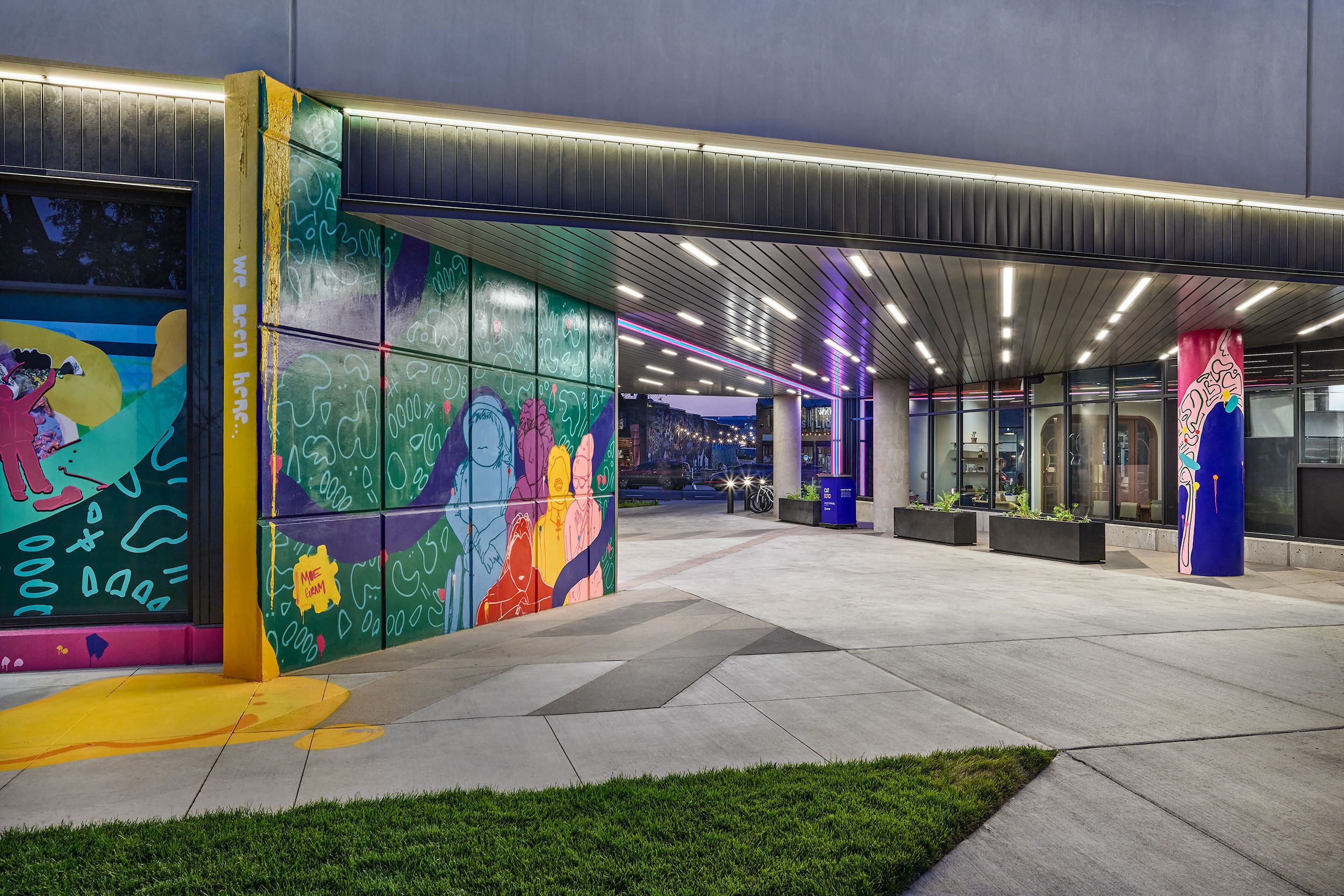 Una entrada moderna a un edificio con murales coloridos, un paseo techado y un área bien iluminada con asientos.