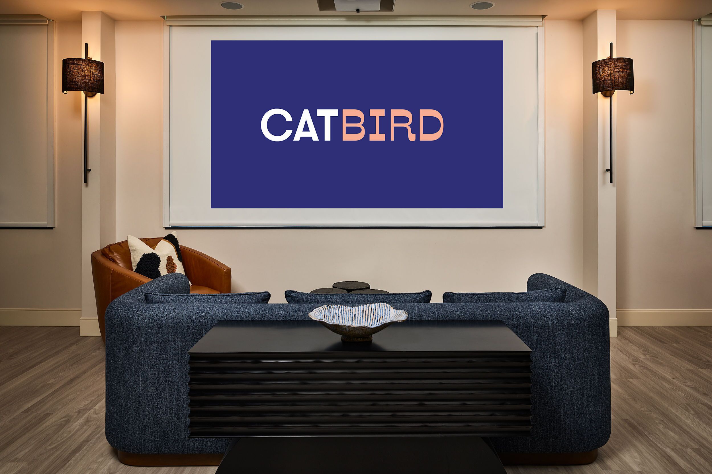 Un salon moderne doté d’un grand écran diffusant l’image de « CATBIRD », d’un canapé bleu, d’une table basse et de deux lampes fixées au mur.