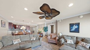A spacious living room with a large ceiling fan, a kitchen in the background, and a dining area.
