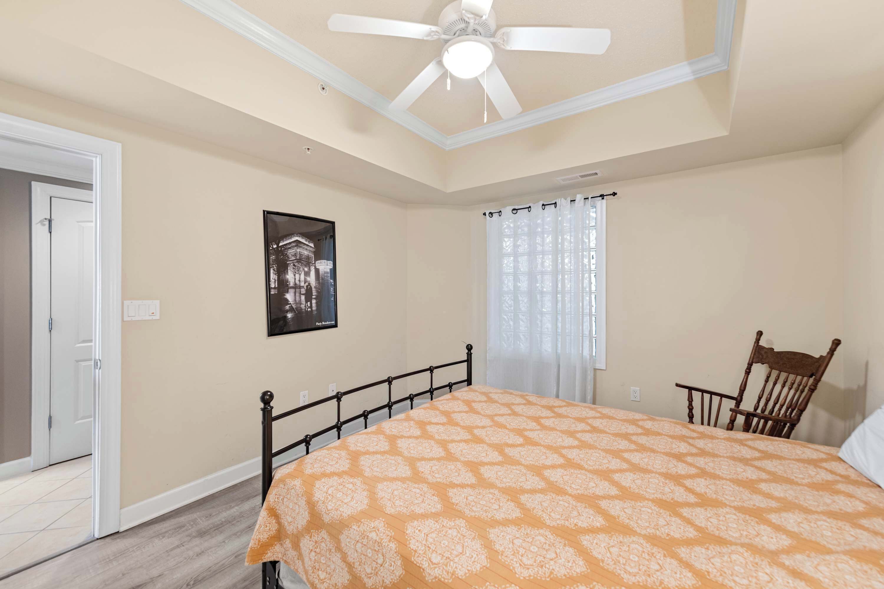 Un dormitorio con cama, ventilador de techo, ventana con cortinas, puerta y un cuadro en la pared.