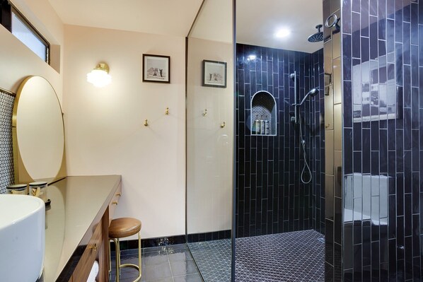 A bathroom with a white vanity, a round mirror, two stools, a shower with black tiles, and a small window with a view.