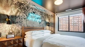 A bedroom with a bed, a nightstand, a wall-mounted lamp, a neon sign, and a window with a decorative screen.