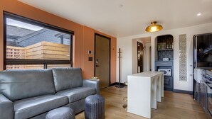 A modern living room with a leather sofa, ottoman, and a small white table. There is a wooden floor, a large window with a view of buildings, and a kitchen area with appliances and decorative tiles.
