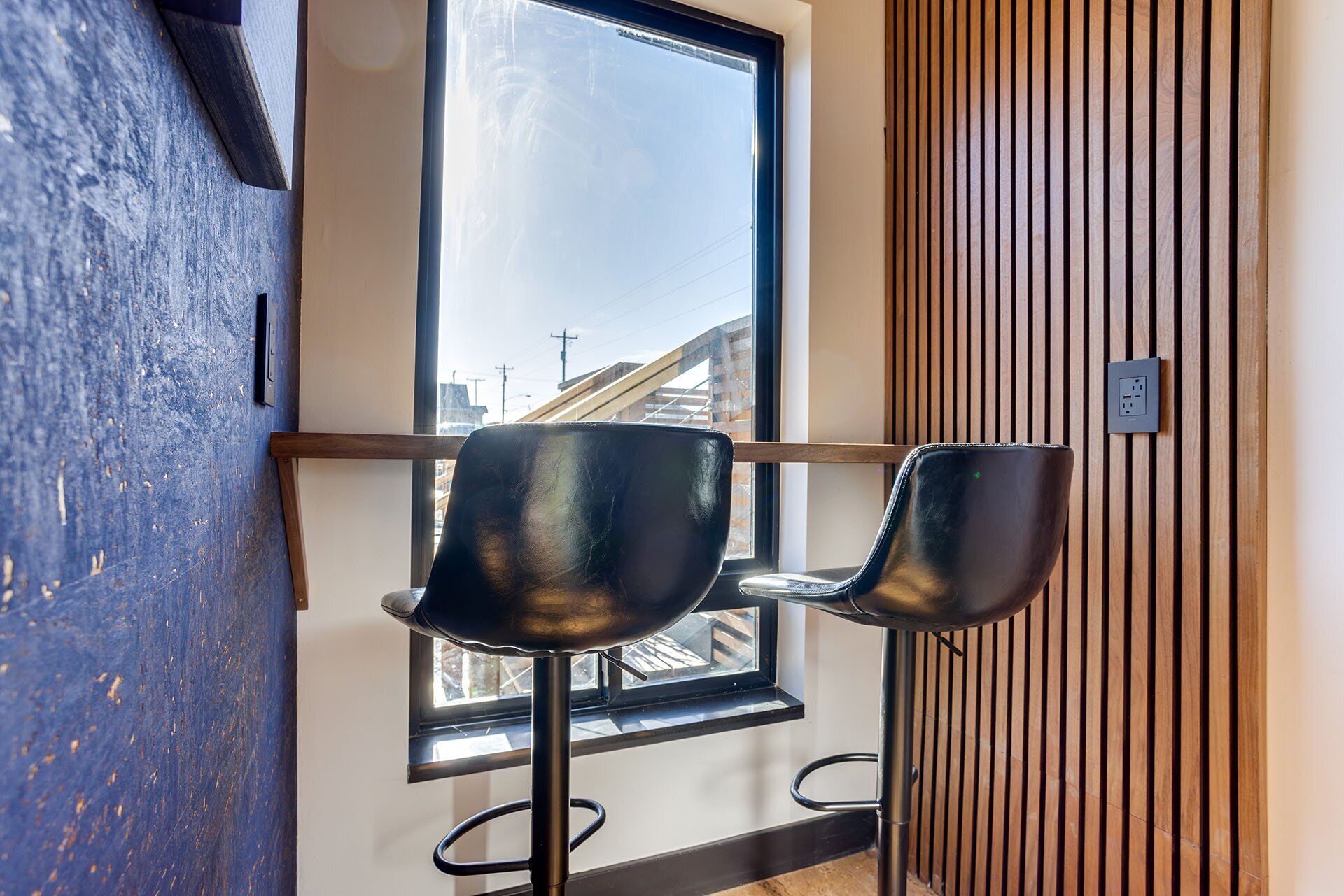 Un interior moderno con dos taburetes negros, un ventanal con vistas a edificios y una pared de listones de madera.