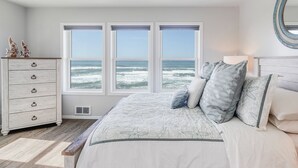 A bedroom with a large bed, a dresser, and a view of the ocean through three windows.