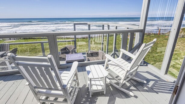 A deck with white Adirondack chairs and a small table, overlooking a beach with the ocean in the background.