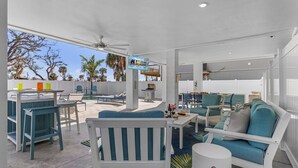 Un espace extérieur aménagé avec des meubles blancs et bleus, protégé par un auvent, doté d’un ventilateur de plafond et d’une enseigne indiquant « Roelens Vacations ».