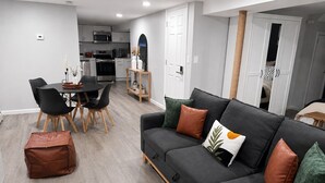 A modern living room with a grey sofa, a dining area with a round table and chairs, and a kitchen in the background. - Boutique Studio 10 min. walk to UBS Arena (Elmont)