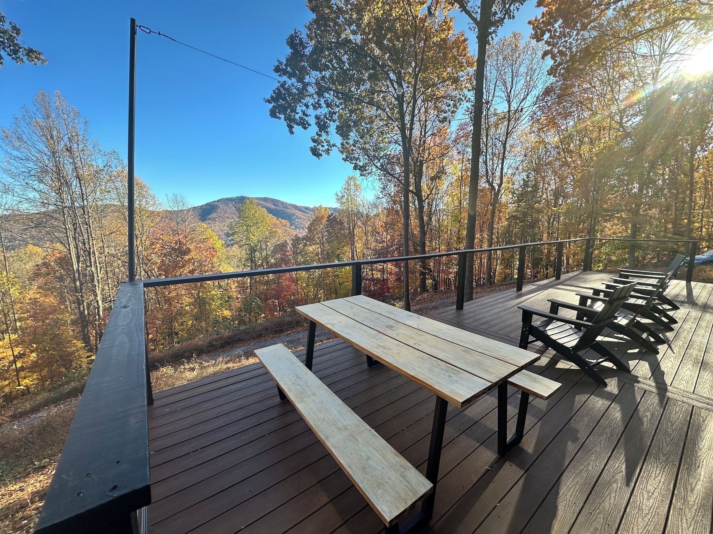 A wooden deck with picnic tables and benches, overlooking a forested area with autumn foliage and distant mountains.