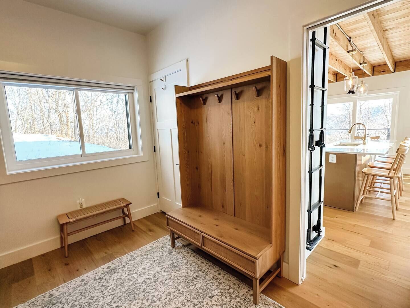 A room with a wooden bench, a large window, and a kitchen area visible through an open door.