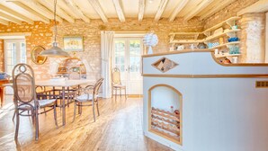 Una cocina rústica con paredes de piedra, vigas de madera, un comedor con sillas de mimbre y una isla blanca con estantes.