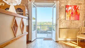 Una habitación con suelo de madera, un radiador blanco, una silla de madera y un balcón con vistas al campo.