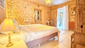 Un dormitorio con pared de piedra, una cama con colcha a rayas, una silla de madera y un escritorio con lámpara.