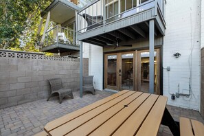 Eine Terrasse mit einem Holztisch und zwei Korbstühlen, ein Balkon mit einem Korbstuhl und eine weiße Ziegelwand.