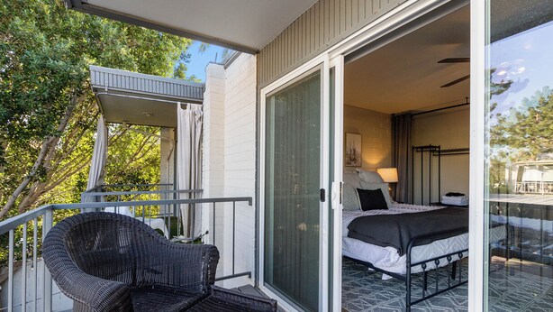 Ein Balkon mit Korbstuhl, ein Bett mit schwarzem Kopfteil und eine Glastür zu einem Zimmer.