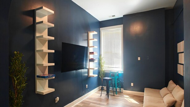 A modern living room with a dark blue wall, a wooden floor, a beige sofa, a TV mounted on the wall, and a window with blinds. - I Street Capsule Hostel (Washington)
