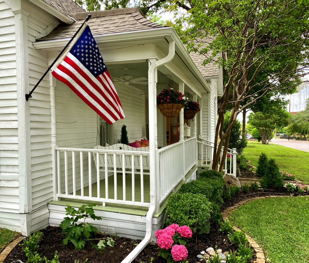 Een wit huis met een veranda, een Amerikaanse vlag en een tuin met roze bloemen.