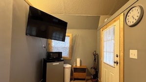 Un couloir étroit avec un téléviseur fixé au mur, un petit réfrigérateur, une poubelle et une horloge sur la porte.