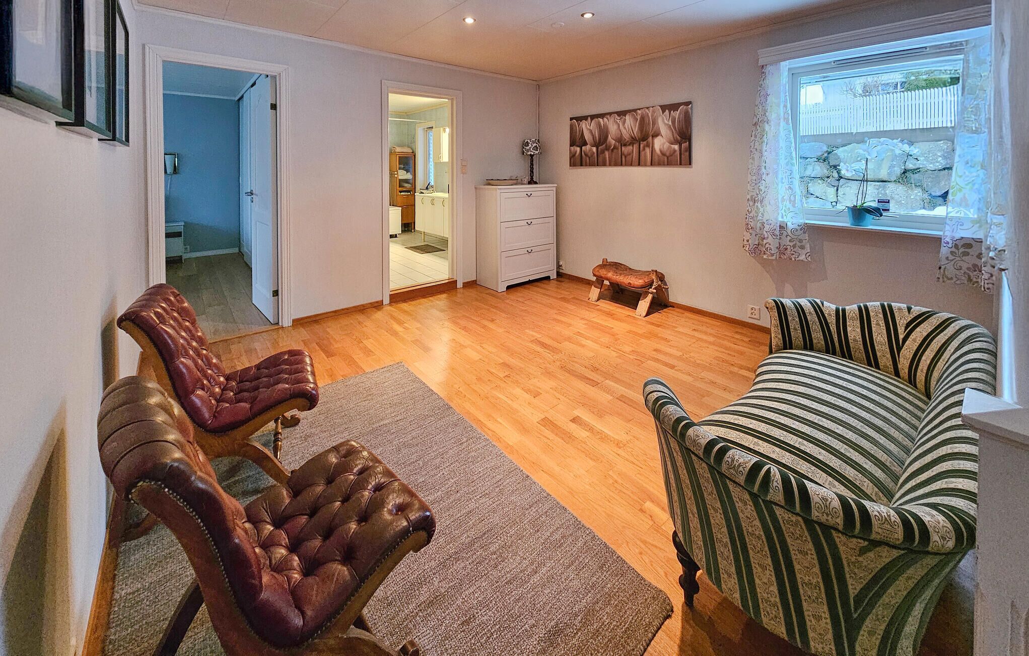 Un salon avec un plancher en bois, deux fauteuils en cuir, un canapé à rayures, un petit tabouret en bois et un tableau accroché au mur.