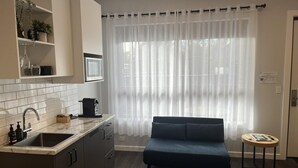 A compact kitchen with a sink, microwave, and cabinets, a dark blue sofa, and a small round table with a book and remote.