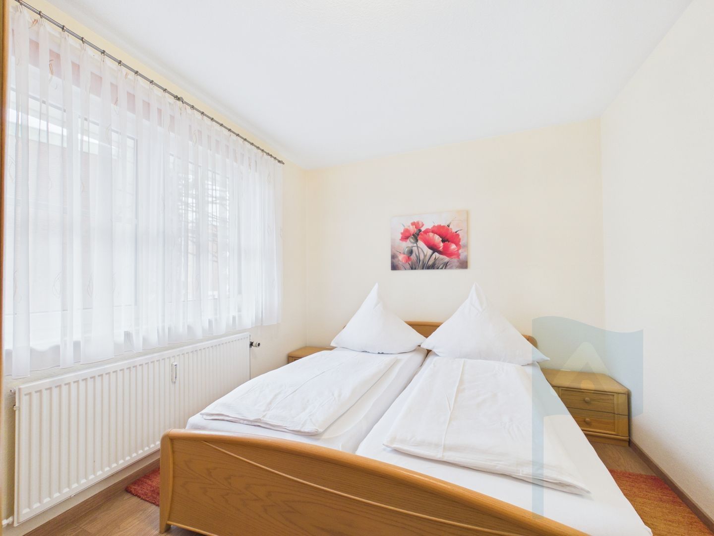 A bedroom with a bed, a radiator, a window with sheer curtains, a bedside table, and a wall-mounted painting.