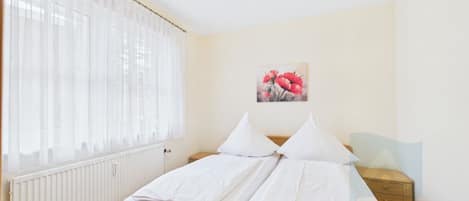 A bedroom with a bed, a radiator, a window with sheer curtains, a bedside table, and a wall-mounted painting.