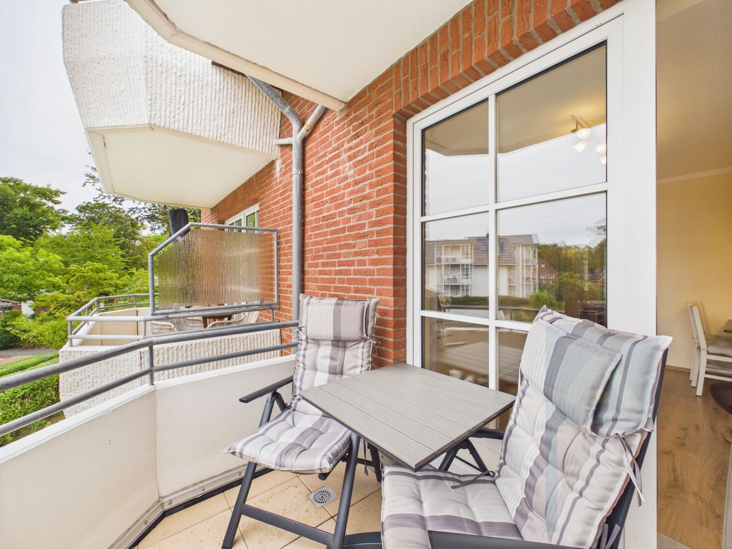 A balcony with a table and chairs, a large window, and a view of greenery and buildings.
