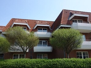 Un edificio residencial de varios pisos con techo de tejas rojas, balcones y un seto en primer plano.