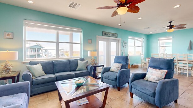 Un salon spacieux avec un canapé bleu, des fauteuils et une table basse. De grandes fenêtres et des ventilateurs de plafond sont présents, ainsi qu’une enseigne indiquant « MAISON DE PLAGE ».