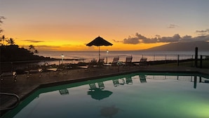 Une piscine avec des chaises longues et un parasol, offrant une vue sur le coucher de soleil sur l’océan.
