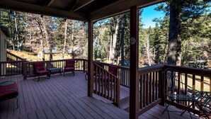 A covered deck with wooden flooring and metal railings, surrounded by trees and a clear sky. - Relaxation Station (Ruidoso)